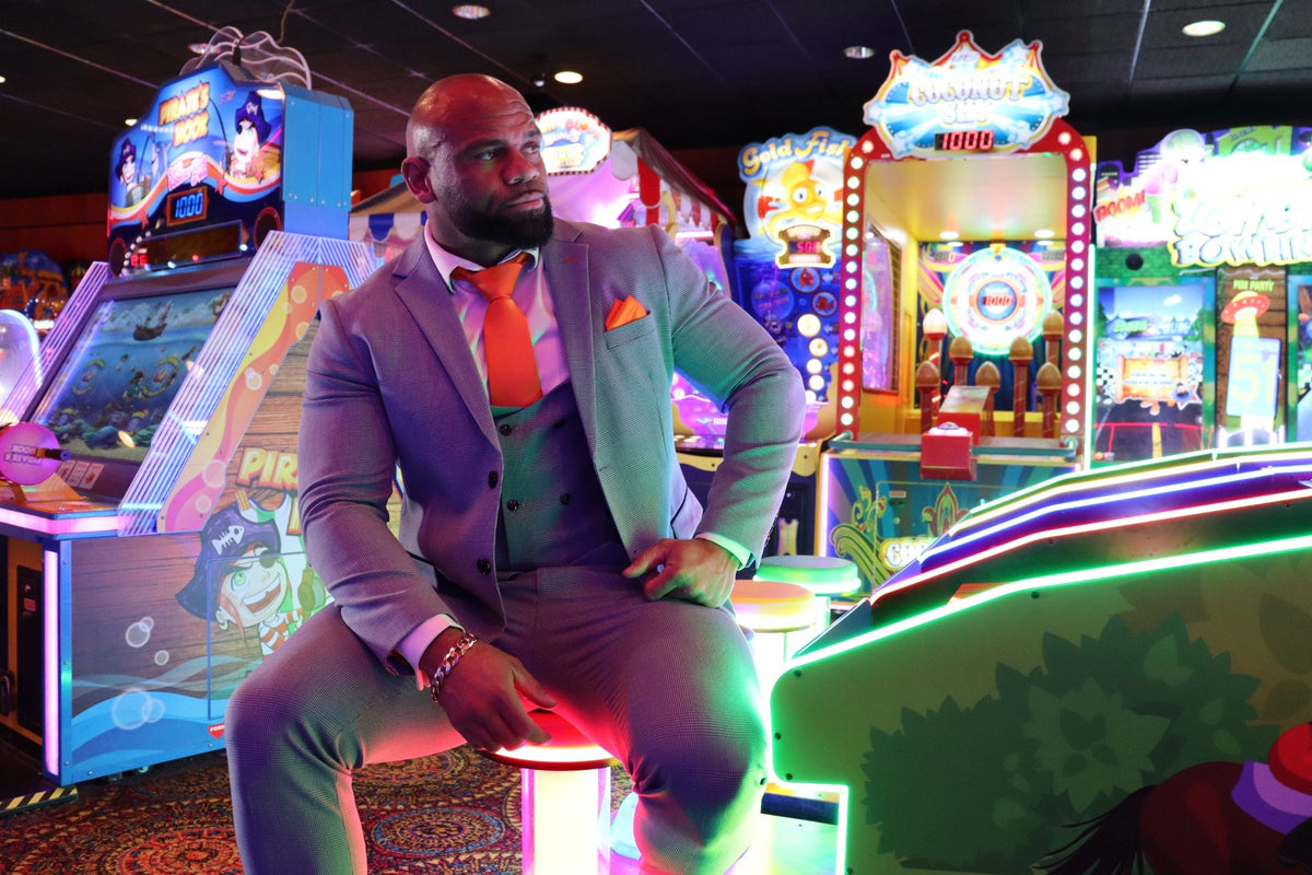 Man wearing the SUITBAE Charles Sky Three-Piece Suit in light blue check with orange tie and pocket square, sitting inside a Blackpool arcade — stylish men’s wedding, prom and race day suit, available in plus sizes.