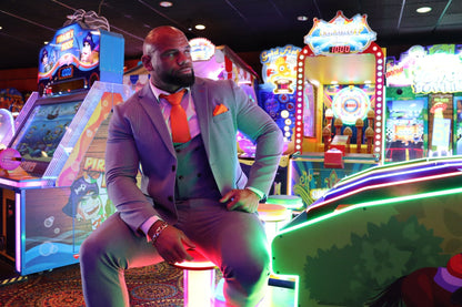 Man wearing the SUITBAE Charles Sky Three-Piece Suit in light blue check with orange tie and pocket square, sitting inside a Blackpool arcade — stylish men’s wedding, prom and race day suit, available in plus sizes.