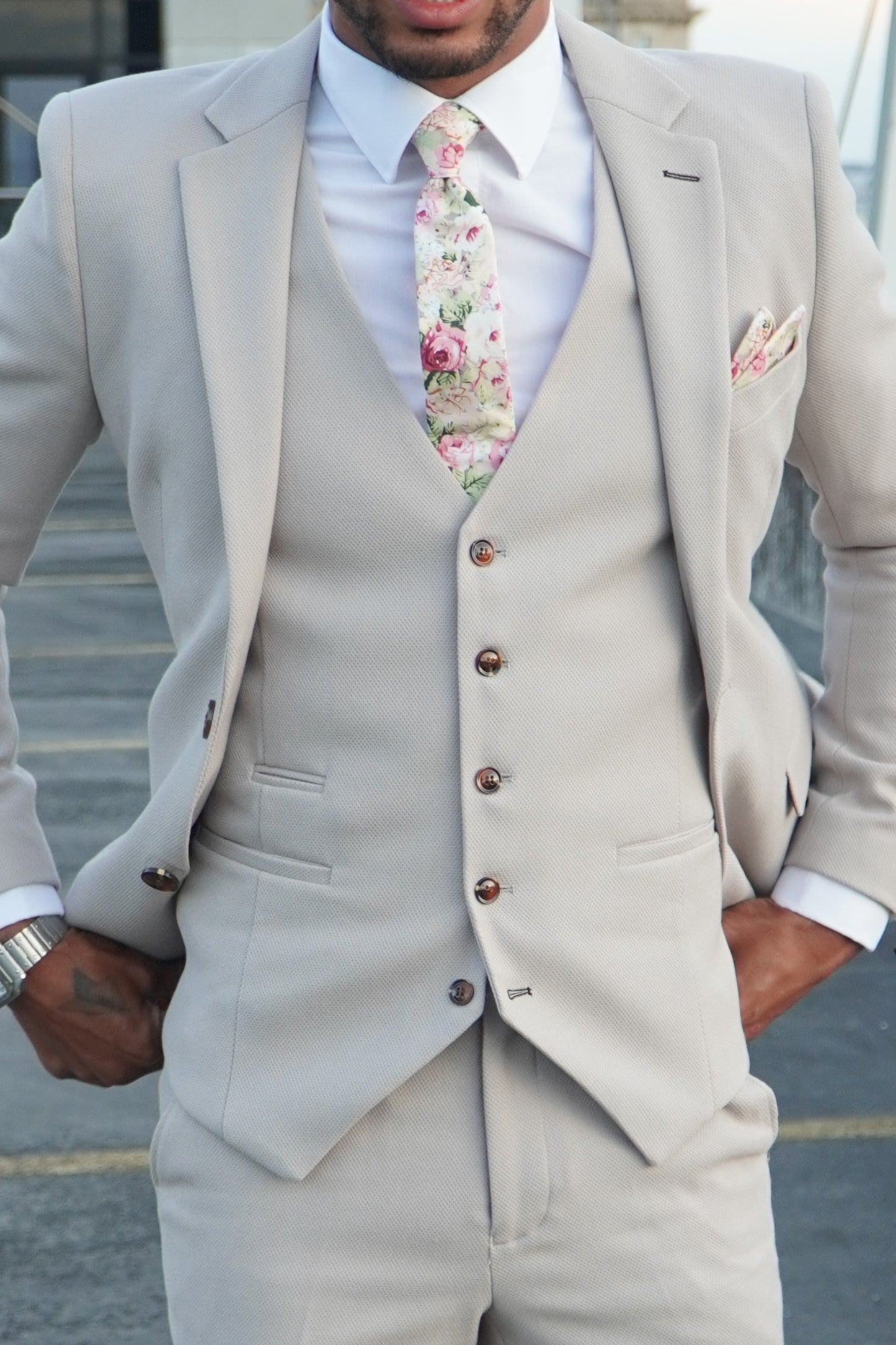 Model wearing the SUITBAE Escobar Stone Three-Piece Suit — tailored beige suit with brown buttons, styled with a floral tie and pocket square, ideal for weddings and summer events.
