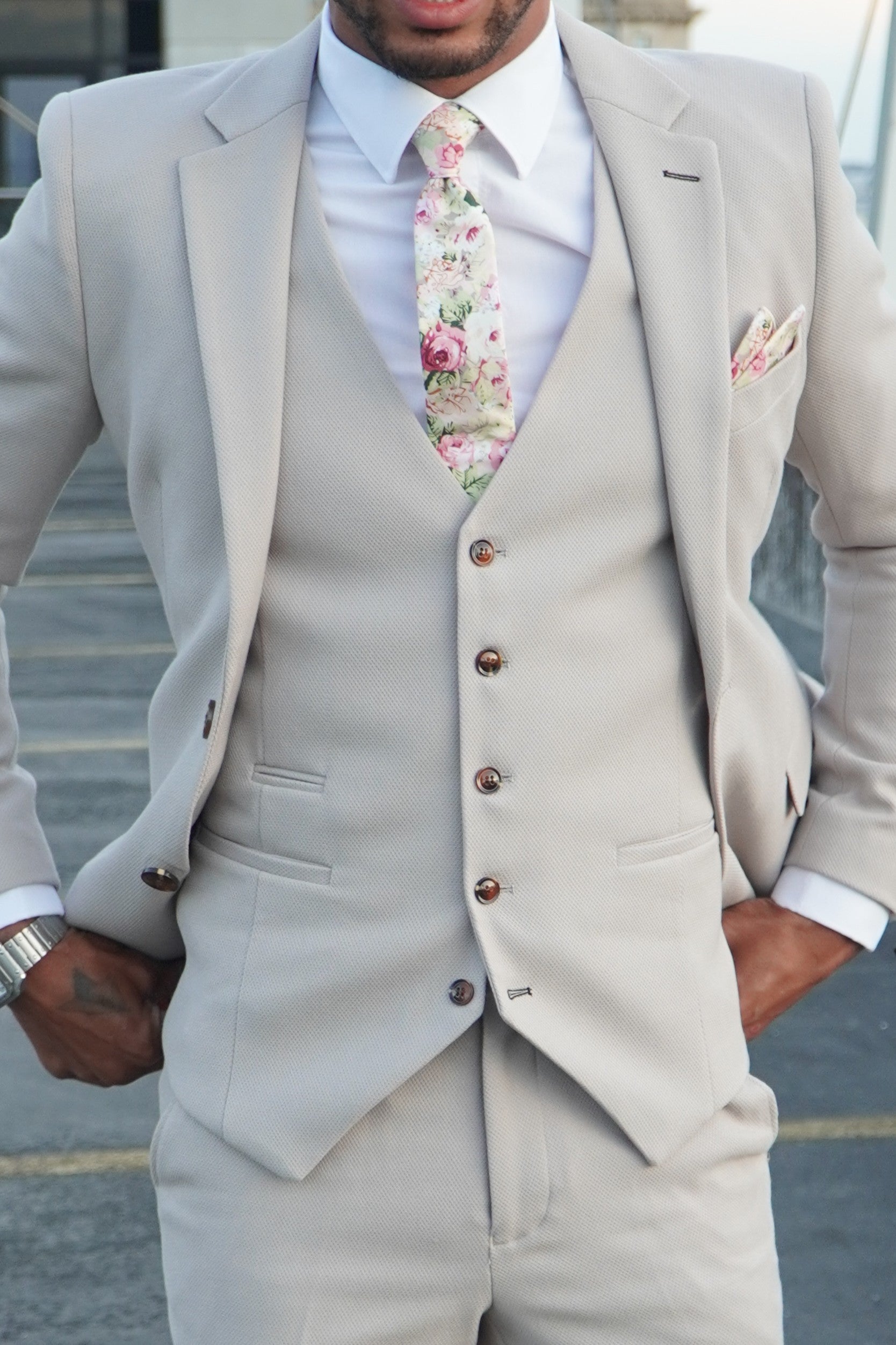Model wearing the SUITBAE Escobar Stone Three-Piece Suit — tailored beige suit with brown buttons, styled with a floral tie and pocket square, ideal for weddings and summer events.
