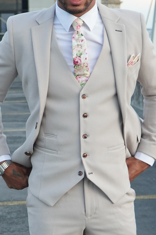 Model wearing the SUITBAE Escobar Stone Three-Piece Suit — tailored beige suit with brown buttons, styled with a floral tie and pocket square, ideal for weddings and summer events.